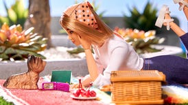 Barbie lying on picnic blanket with a dog, a book, a flip phone and some cherries