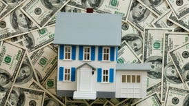 a model of a white house with blue shutters and a gray roof sits atop a large pile of $100 bills all spread around